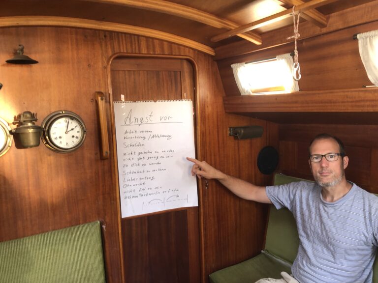 Christian Mentzel ist in der Kajüte und zeigt mit dem Finger auf einen Flipchart, auf dem steht "Angst vor". Er macht Bewusstseinsarbeit. Seine Augen gucken klar und vertrauensvoll in die kamera.