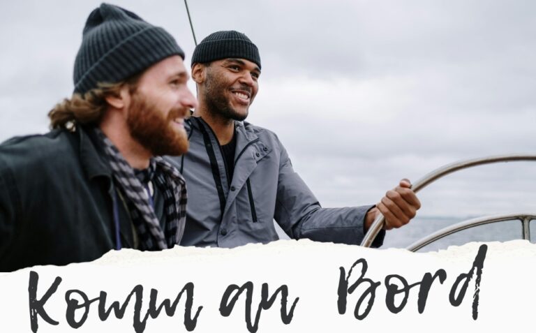 2 Männer sind auf einem Boot, der eine hält sich an einem Griff beim Steuerrad fest. Beide sind in dem Moment, und strahlen im Gesicht.